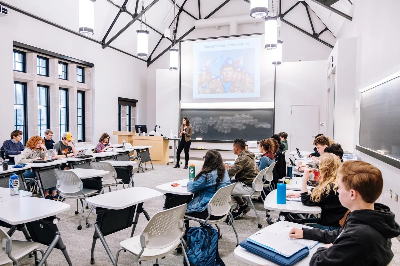 A professor lecturing a full classroom of students seated at desks with laptops, in front of a projected slide titled "Humanitarian Intervention."