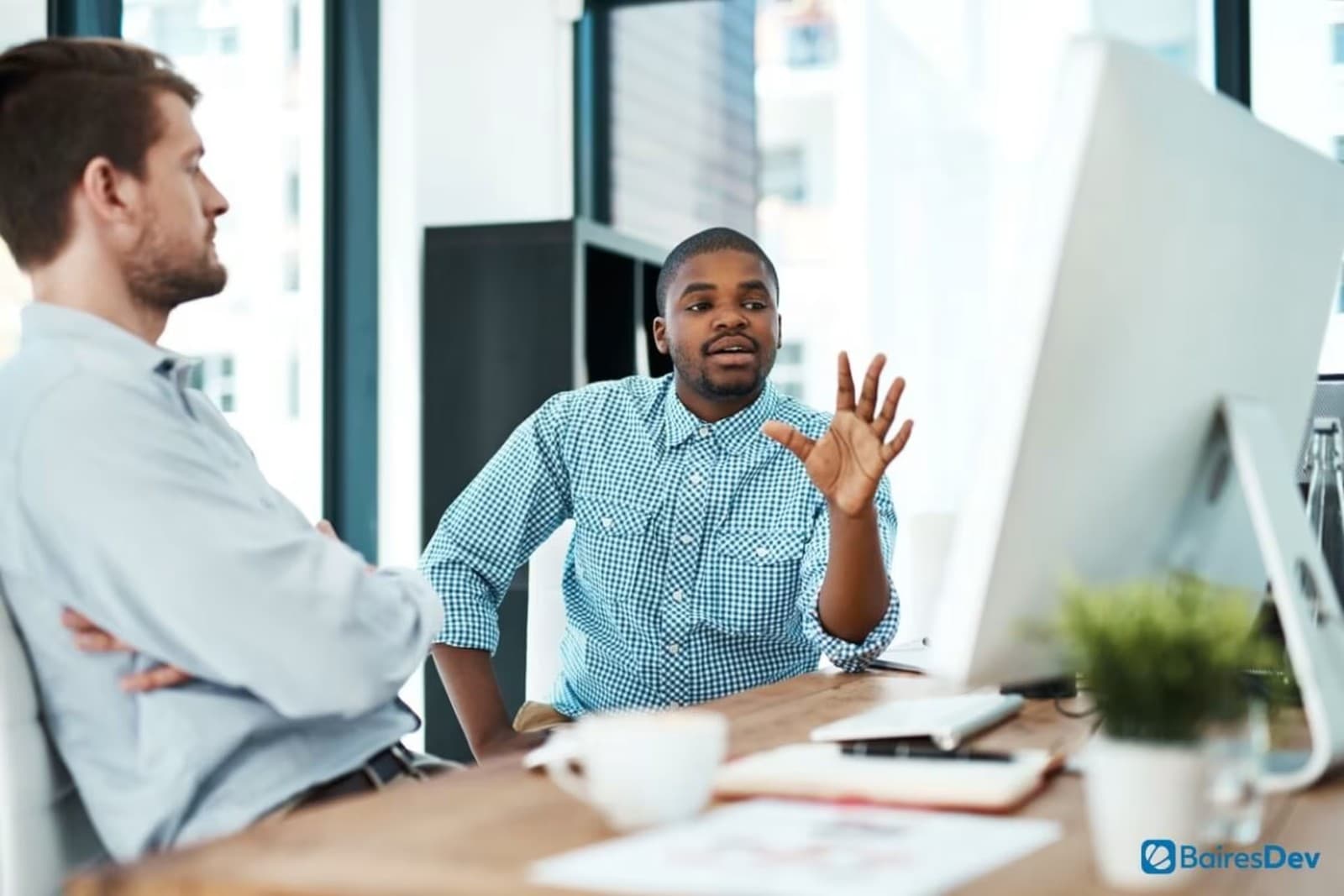 A young man in a checkered shirt gesturing while explaining something to a colleague at a desk with a large computer monitor.