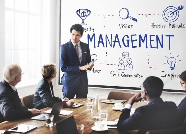 A businessman presenting to seated colleagues in front of a whiteboard illustration labeled "Management" with icons for vision, communication, and leadership.
