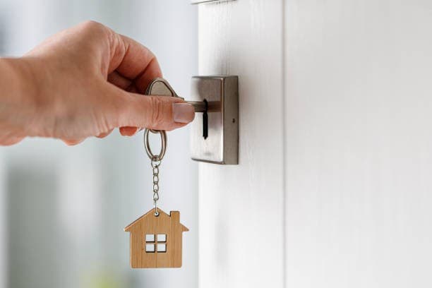 A close-up of a hand inserting a key with a wooden house-shaped keychain into a front door lock.