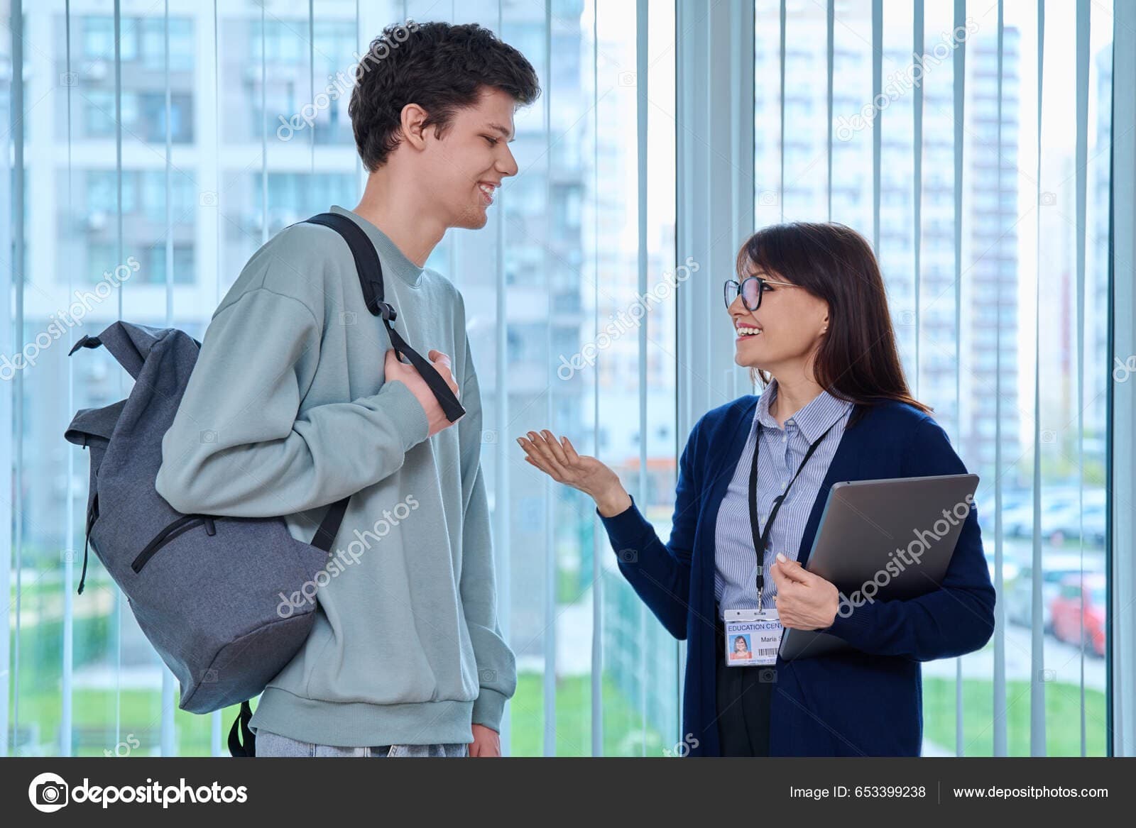 A student wearing a backpack smiling and talking with a smiling advisor holding a laptop in a bright office hallway.