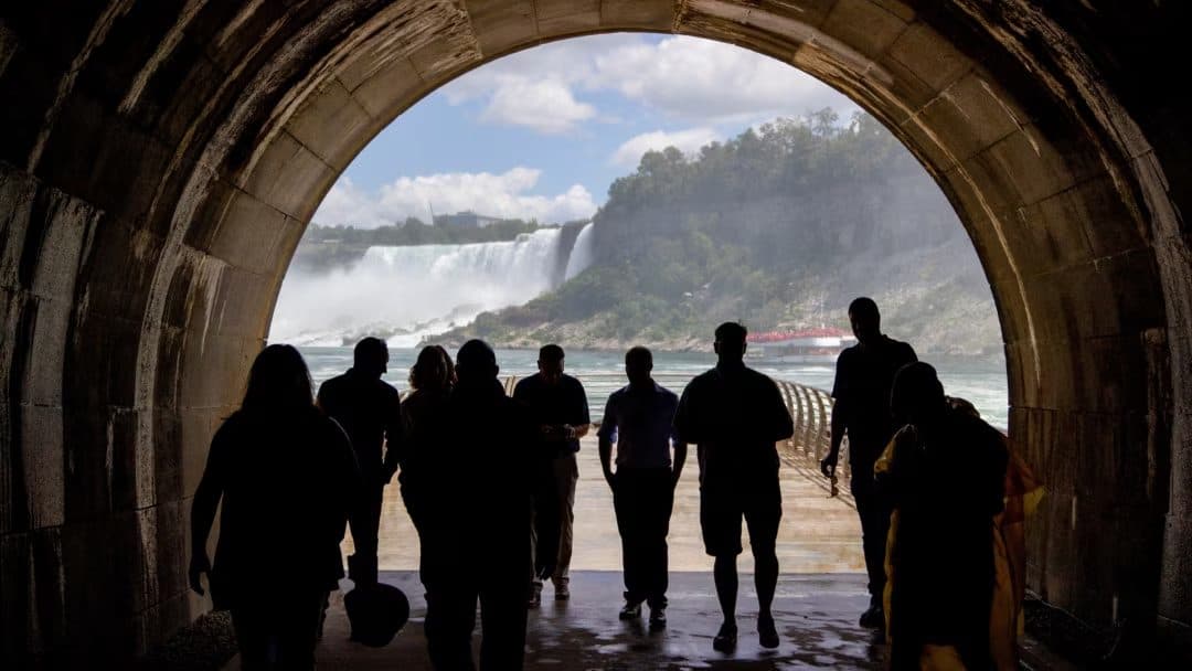 Silhouetted visitors walking through a stone tunnel toward a sunlit view of a waterfall and river gorge.