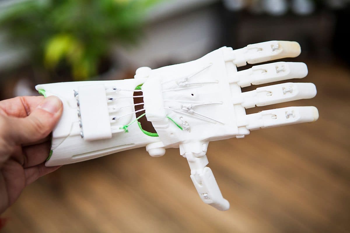 A hand holding a white 3D-printed prosthetic hand with visible tendon strings and jointed, articulated fingers.