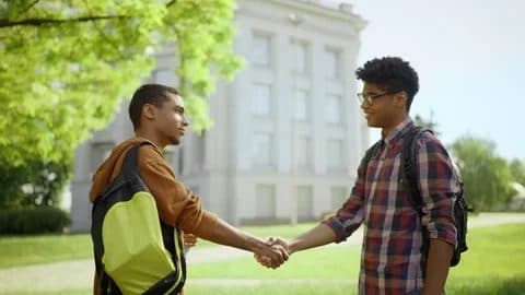 Two young men with backpacks shaking hands outdoors in front of a building, suggesting a mentorship or business meeting.