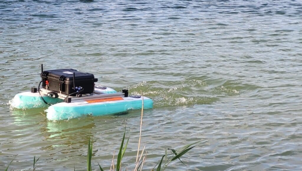 A small teal autonomous rover with a black equipment case floating on open water, leaving a wake near a reed-lined shore.