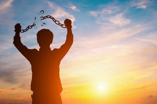 Silhouette of a person at sunset raising broken chains overhead with both arms, symbolizing freedom.