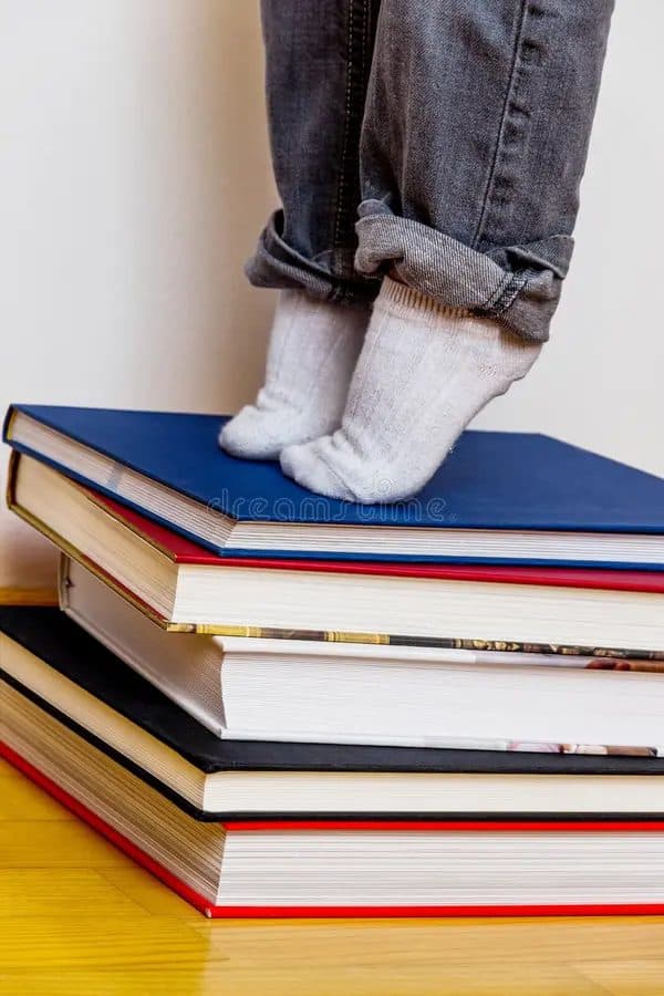 A child in white socks standing on top of a tall stack of hardcover books.