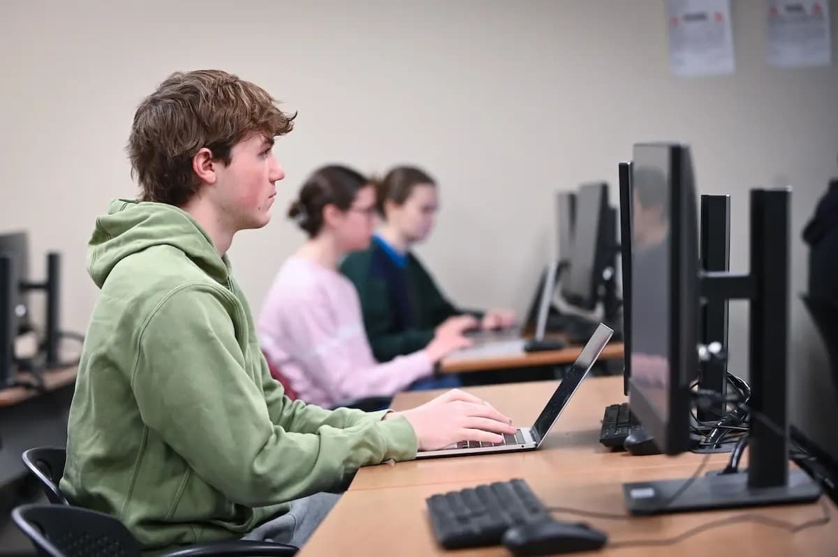 A student in a green hoodie works on a laptop at a row of computer workstations, with classmates studying at monitors behind him.