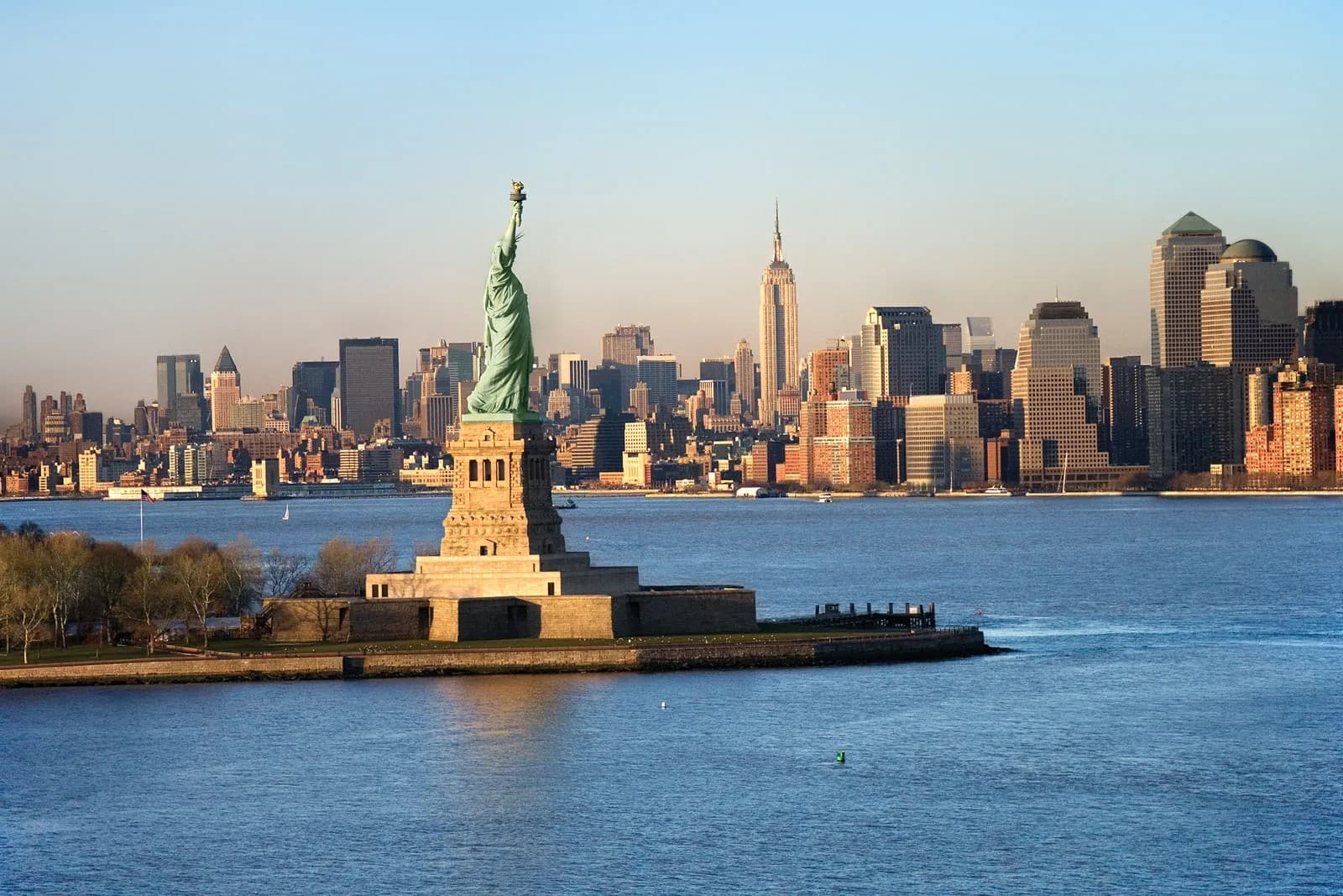 The Statue of Liberty at sunset with the Manhattan skyline, including the Empire State Building, rising behind New York Harbor.