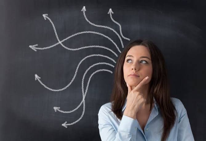 A woman looks upward thoughtfully in front of a chalkboard with hand-drawn arrows branching outward, representing many possible directions.