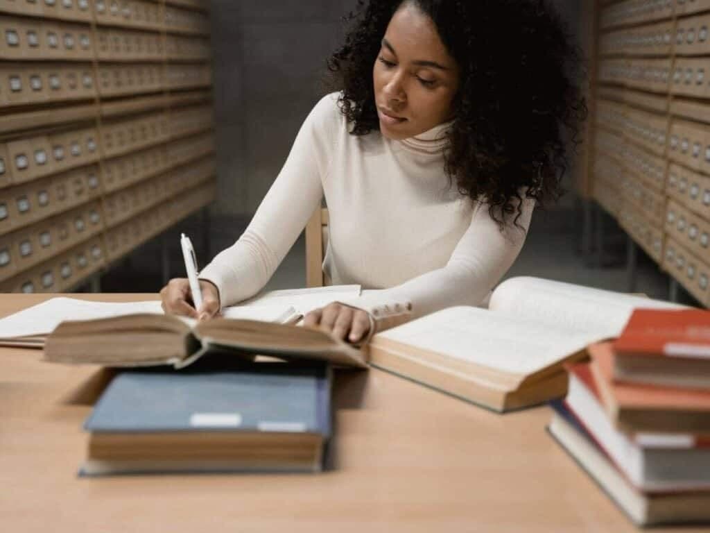 A student in a library annex writes notes while reading a large open book, surrounded by card-catalog cabinets and stacks of books.
