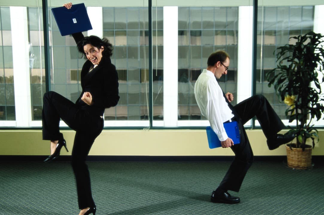 Two office workers in business attire joyfully kicking up their heels in celebration, holding folders in a bright office.