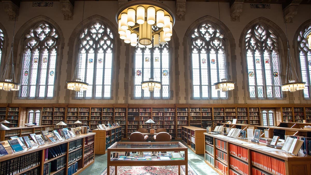 A Gothic-style reading room in a Yale library, with tall stained-glass windows, wood bookshelves, and hanging chandeliers.