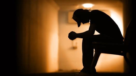 The silhouette of a person sitting alone with their head down and arms resting on their knees in a dim, glowing hallway.