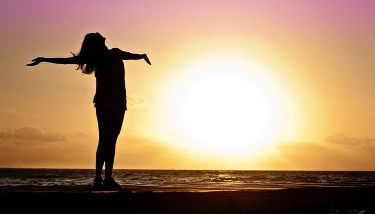 Silhouette of a woman on a pier at sunset, arms outstretched toward a bright sun over the ocean.