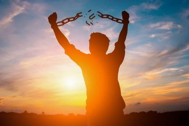 Silhouette of a person raising both arms as a chain around their wrists breaks apart, set against a sunset sky.